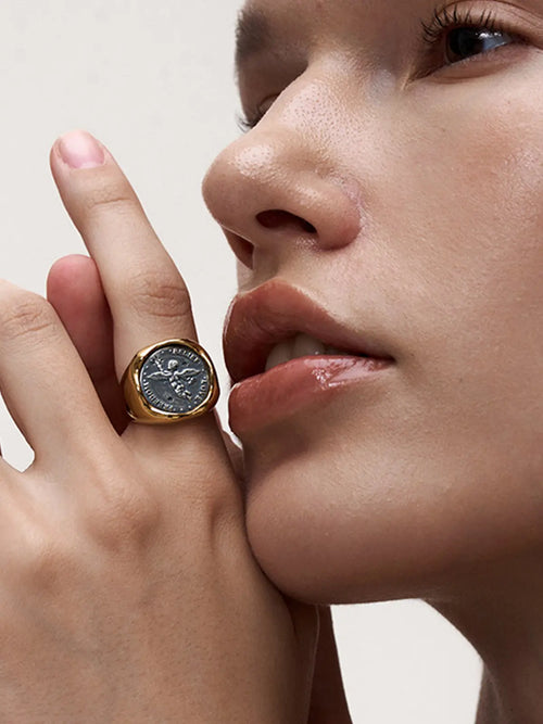 Model wearing the Tactilery Angel Coin Ring on the index finger with hand raised near face, the large polished gold round coin medallion with engraved classical angel relief and rope-twist border facing forward, against a warm beige background wearing a dark brown sleeveless top