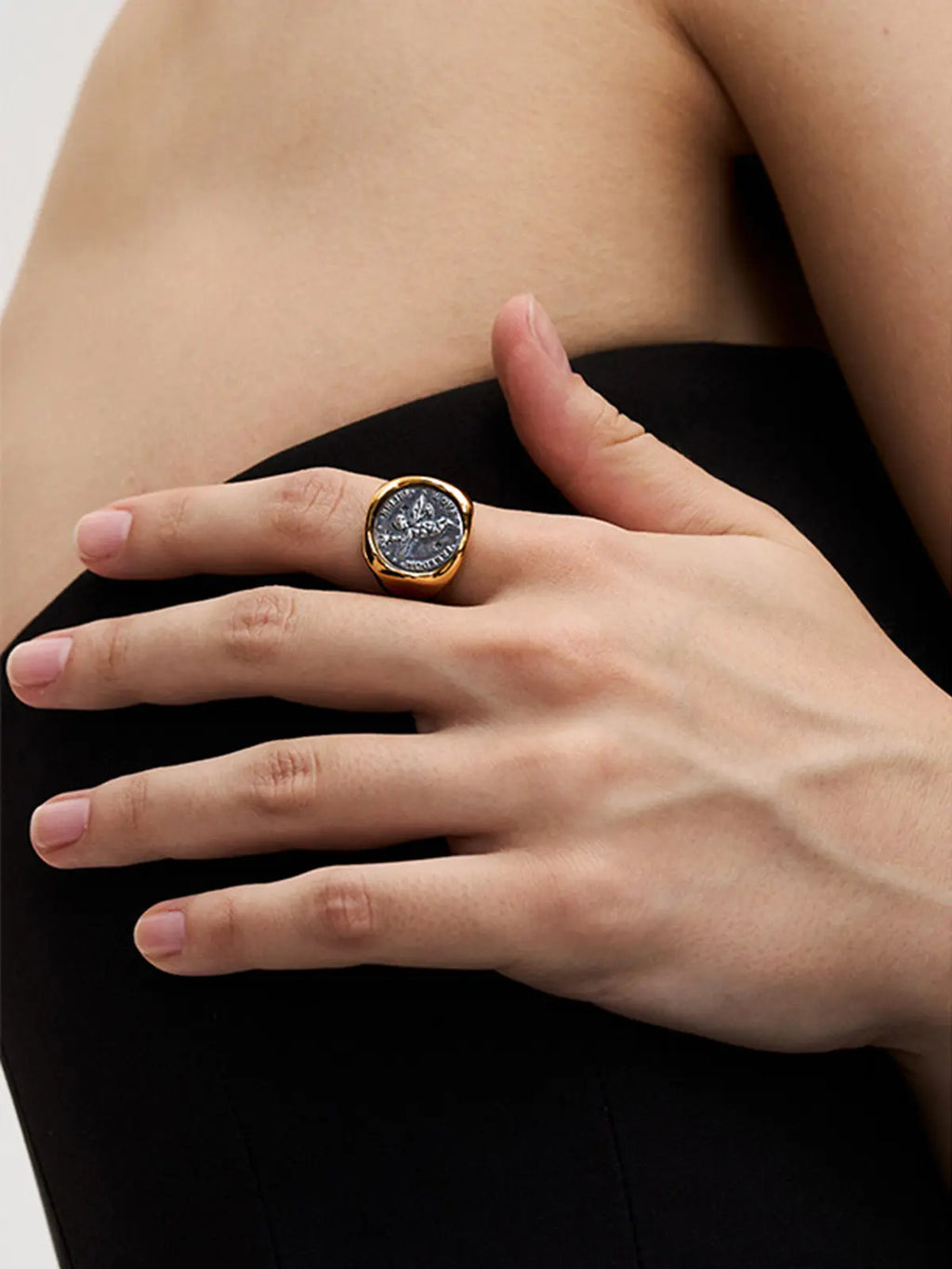 Close-up of a model's hand wearing the Tactilery Angel Coin Ring on the index finger against a dark background, the large polished gold round coin medallion with engraved classical angel relief and rope-twist border centered on the finger