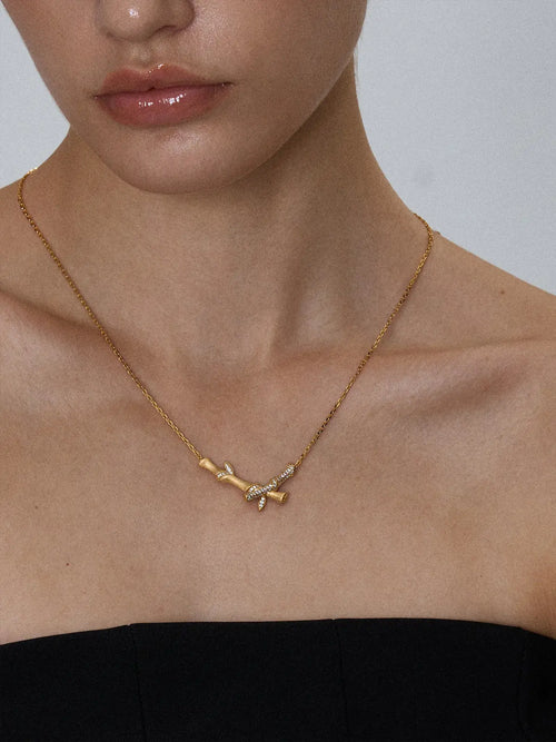 Close-up of a model's décolletage wearing the Tactilery Bamboo Whisper Necklace, the three hollow cylindrical bamboo-segment tubes of graduating height in polished gold hanging at the neckline on a fine polished gold twisted rope chain, against a warm taupe background wearing a dark draped top