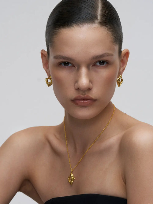 Front-facing editorial portrait of a model wearing the Tactilery Baroque Frame Stud Earrings on both ears, the angular diamond-shaped gold C-hoop with heavily textured molten baroque surface and pavé zircon quatrefoil accent at the lobe, paired with a matching baroque gold pendant necklace on a fine gold chain, against a light grey background wearing a black strapless top with hair slicked back