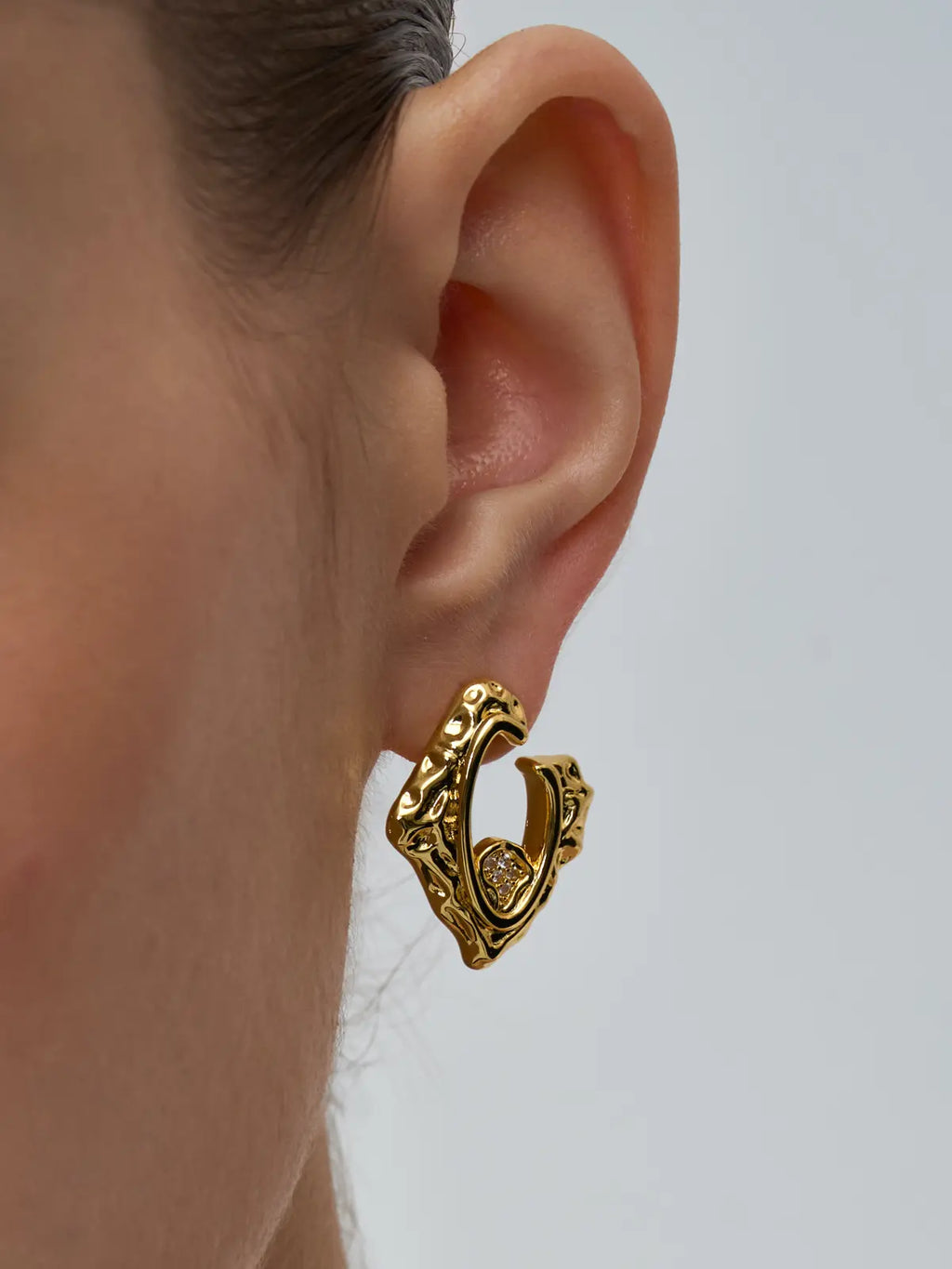 Extreme close-up of a model's ear wearing the Tactilery Baroque Frame Stud Earrings, the angular diamond-shaped gold C-hoop with heavily textured molten baroque surface and open center frame filling the frame, the small pavé zircon quatrefoil accent at the lower inner corner in sharp detail, against a light grey background