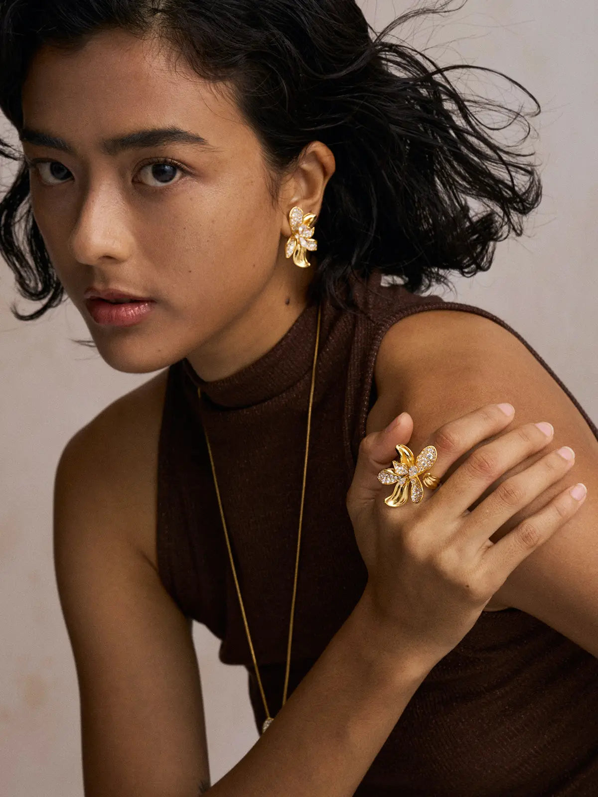 Editorial portrait of a model wearing the Tactilery Blossom Ring on the index finger with hand resting on shoulder, the oversized gold five-petal orchid flower with pavé zircon upper petals and smooth gold lower petals visible, paired with matching Blossom Hoop Earrings and a fine gold chain necklace, against a warm beige background wearing a dark brown sleeveless turtleneck