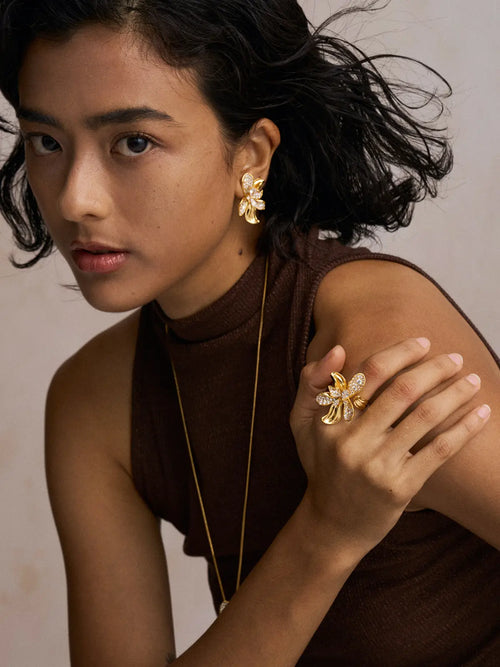 Editorial portrait of a model wearing the Tactilery Blossom Ring on the index finger with hand resting on shoulder, the oversized gold five-petal orchid flower with pavé zircon upper petals and smooth gold lower petals visible, paired with matching Blossom Hoop Earrings and a fine gold chain necklace, against a warm beige background wearing a dark brown sleeveless turtleneck