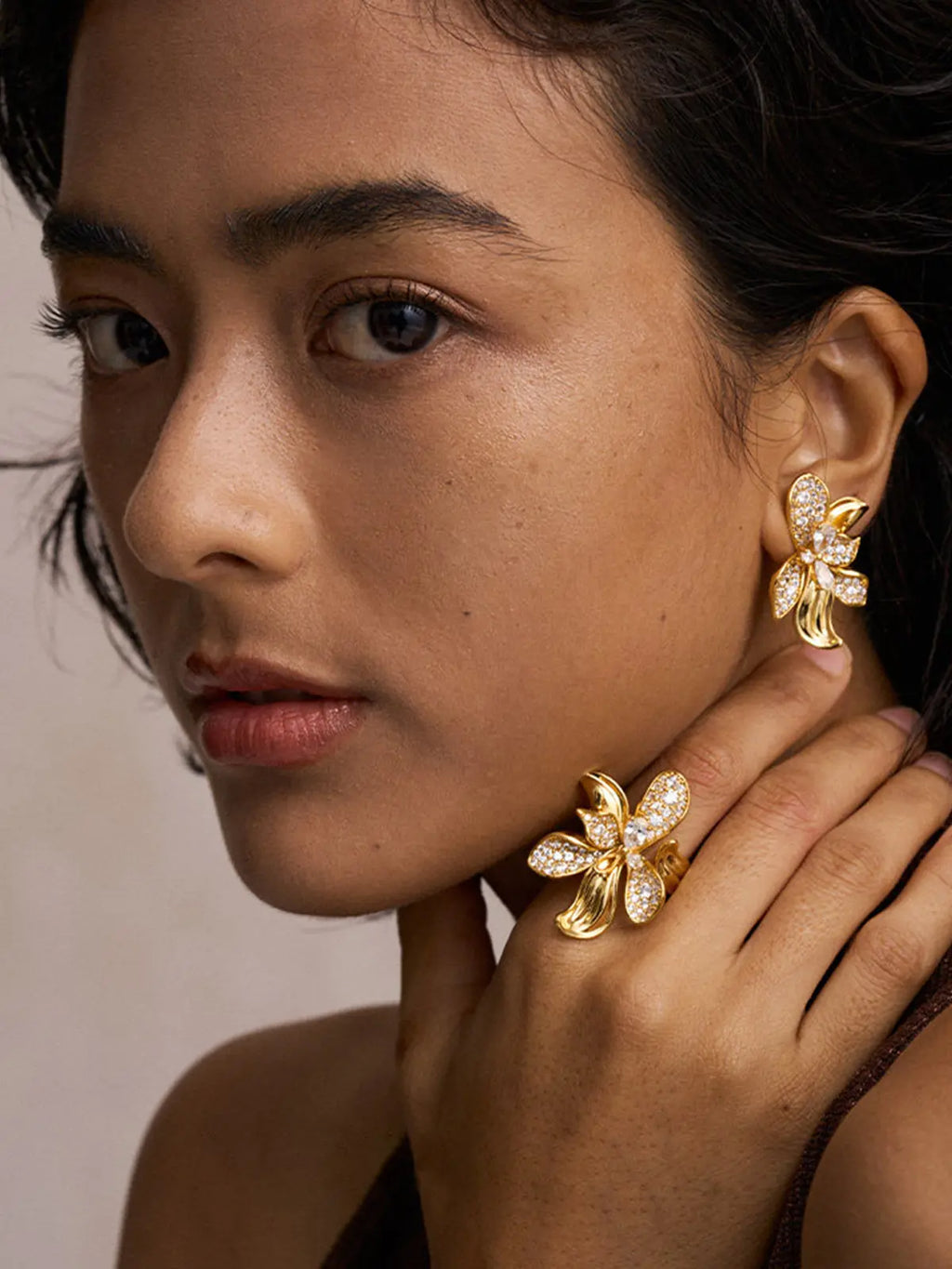 Close-up portrait of a model with hand raised near neck wearing the Tactilery Blossom Ring on the index finger, the oversized gold five-petal orchid flower with three fully pavé zircon upper petals and two smooth gold lower petals with ribbed vein texture visible, paired with matching Blossom Hoop Earrings, against a warm beige background