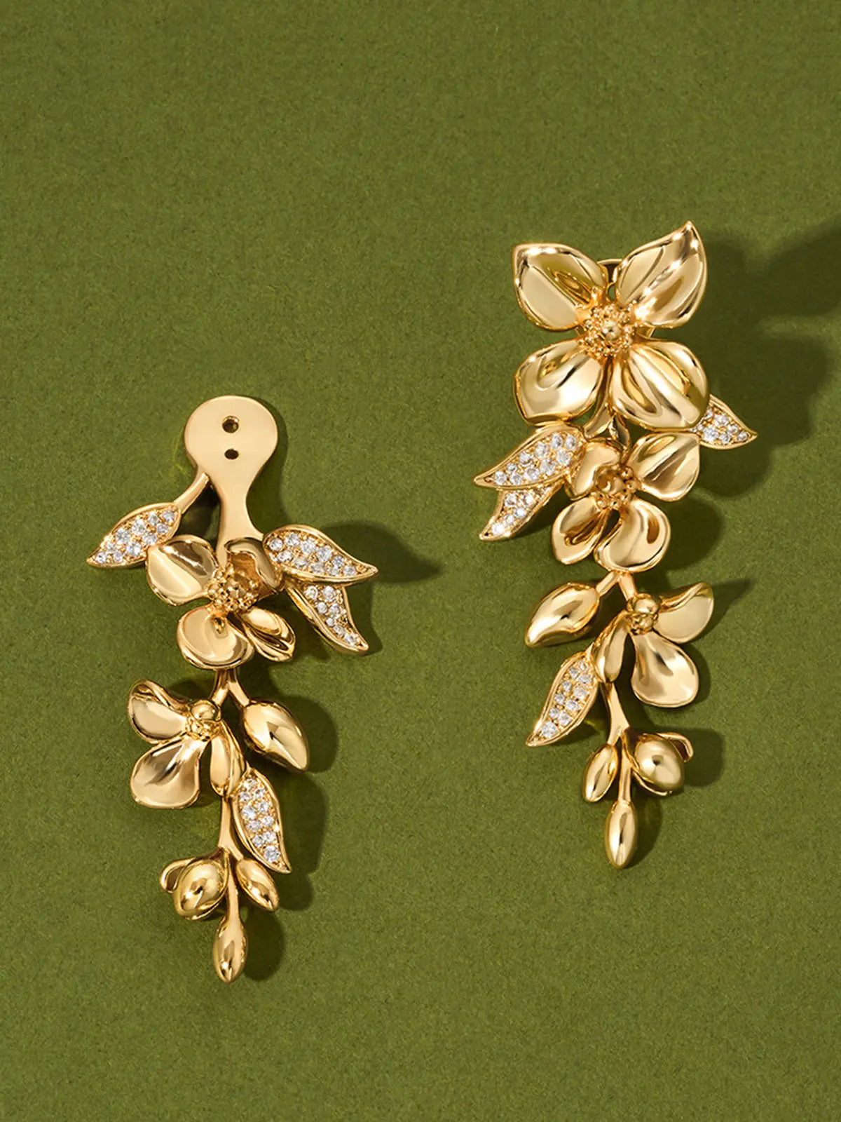 Pair of Tactilery Tassel Detachable Drop Earrings in polished gold displayed on a dark olive green surface, one showing the back of the assembled drop with the gold disc butterfly back and the detachable connector visible, one showing the front of the assembled drop with the large five-petal blossom flower stud top and the smaller blossom flower with trailing polished gold oval leaf bud spray and pavé zircon leaf accents below