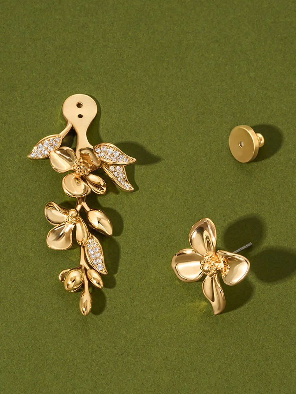 Tactilery Tassel Detachable Drop Earring in polished gold displayed on a dark olive green surface showing the two detachable components separated — the large five-petal blossom flower stud top with smooth domed petals and pavé zircon stamens on the left with the gold disc butterfly back removed, and the smaller five-petal blossom flower with trailing polished gold oval leaf bud spray and pavé zircon leaf accents on the right