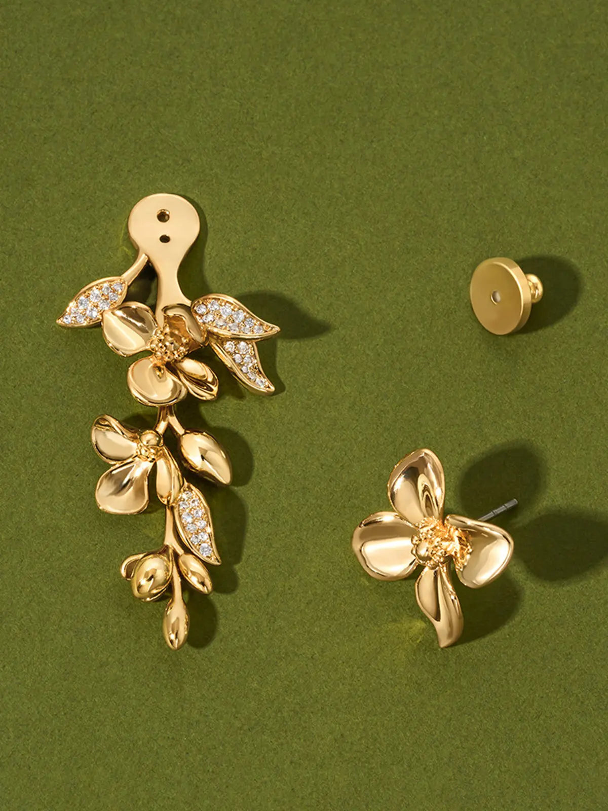Tactilery Tassel Detachable Drop Earring in polished gold displayed on a dark olive green surface showing the two detachable components separated — the large five-petal blossom flower stud top with smooth domed petals and pavé zircon stamens on the left with the gold disc butterfly back removed, and the smaller five-petal blossom flower with trailing polished gold oval leaf bud spray and pavé zircon leaf accents on the right