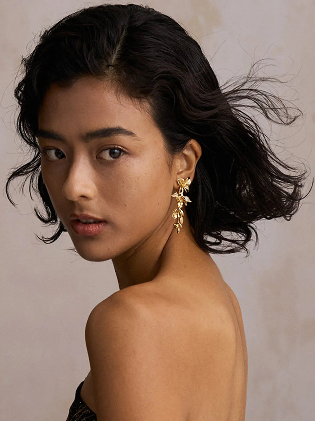 Three-quarter portrait of a model wearing the Tactilery Tassel Detachable Drop Earrings assembled as a full drop on one ear, the long cascading polished gold blossom flower and leaf bud spray dangling from the lobe, against a warm taupe background wearing a dark strapless top with short dark wavy hair