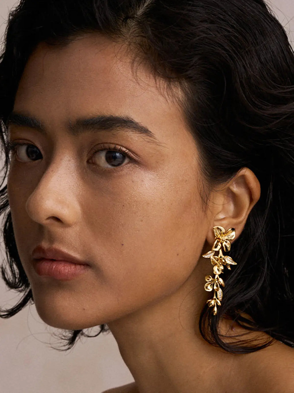Close-up three-quarter portrait of a model wearing the Tactilery Tassel Detachable Drop Earrings assembled as a full drop on one ear, the long cascading polished gold blossom flower and leaf bud spray with pavé zircon leaf accents filling the frame at the lobe, against a warm taupe background wearing a dark strapless top