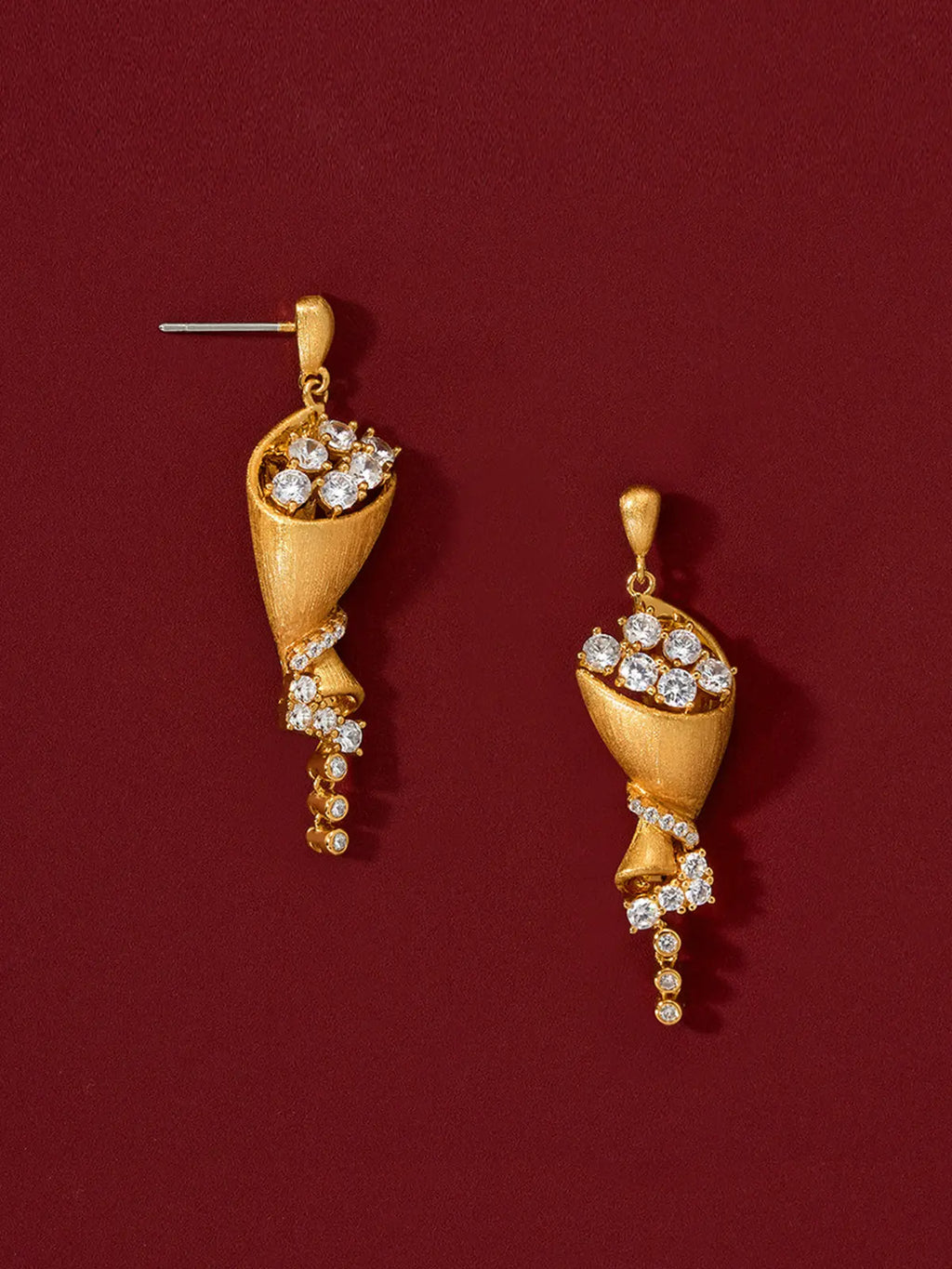 Pair of Tactilery Bouquet Stud Earrings in matte brushed gold displayed upright on a deep red surface, one showing the full front face of the sculptural twisted cone bouquet drop with large round zircon bloom cluster at top, pavé zircon spiral ribbon mid-body, and bezel-set zircon chain tail, one showing the three-dimensional depth of the twisted cone form, with silver post pins visible at top