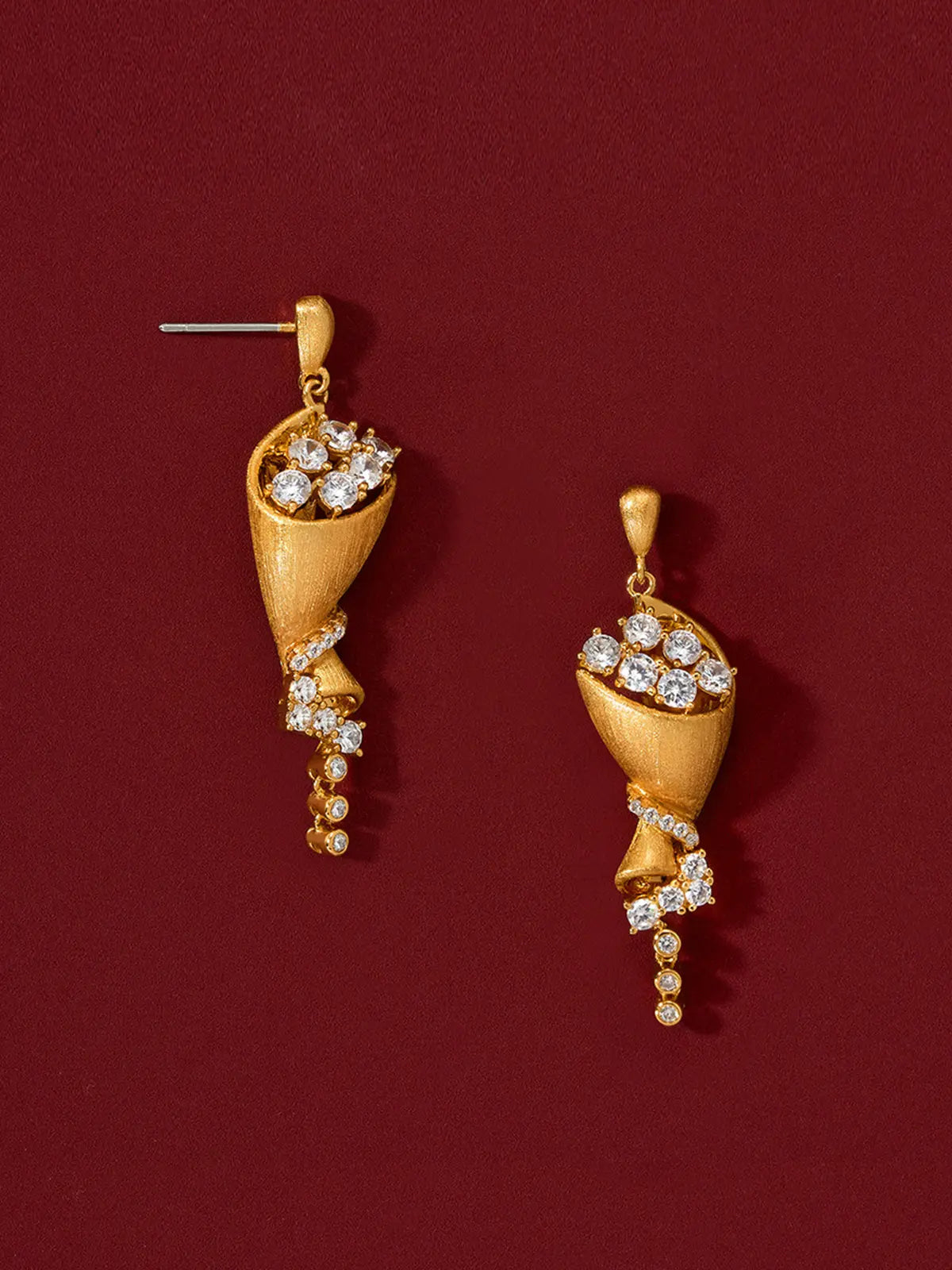 Pair of Tactilery Bouquet Stud Earrings in matte brushed gold displayed upright on a deep red surface, one showing the full front face of the sculptural twisted cone bouquet drop with large round zircon bloom cluster at top, pavé zircon spiral ribbon mid-body, and bezel-set zircon chain tail, one showing the three-dimensional depth of the twisted cone form, with silver post pins visible at top
