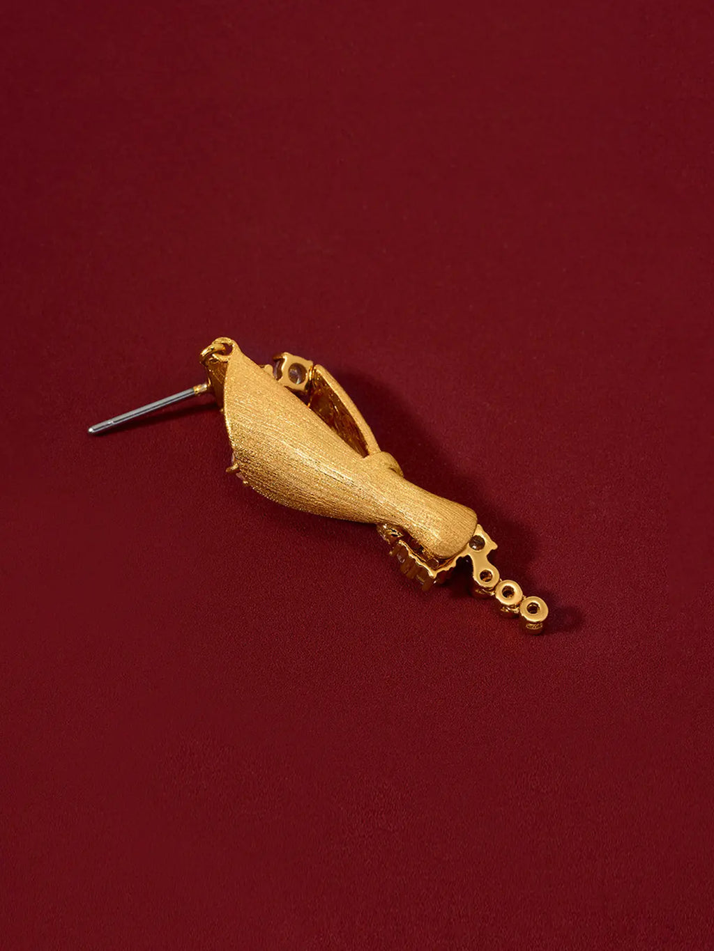 Single Tactilery Bouquet Stud Earring in matte brushed gold displayed lying on its back on a deep red surface, showing the reverse of the sculptural twisted cone bouquet body — the matte brushed gold sand-textured back surface of the cone wrap, the bezel-set zircon chain tail, and the silver post pin at the smooth gold teardrop stud top