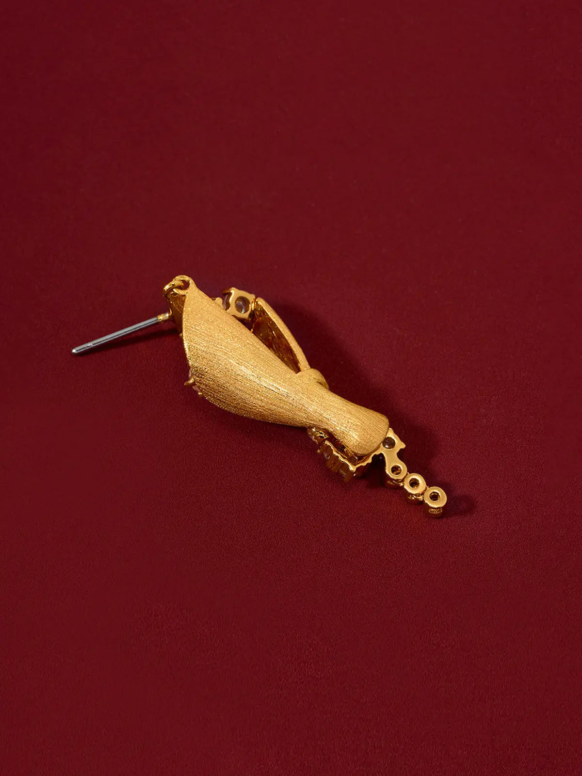 Single Tactilery Bouquet Stud Earring in matte brushed gold displayed lying on its back on a deep red surface, showing the reverse of the sculptural twisted cone bouquet body — the matte brushed gold sand-textured back surface of the cone wrap, the bezel-set zircon chain tail, and the silver post pin at the smooth gold teardrop stud top