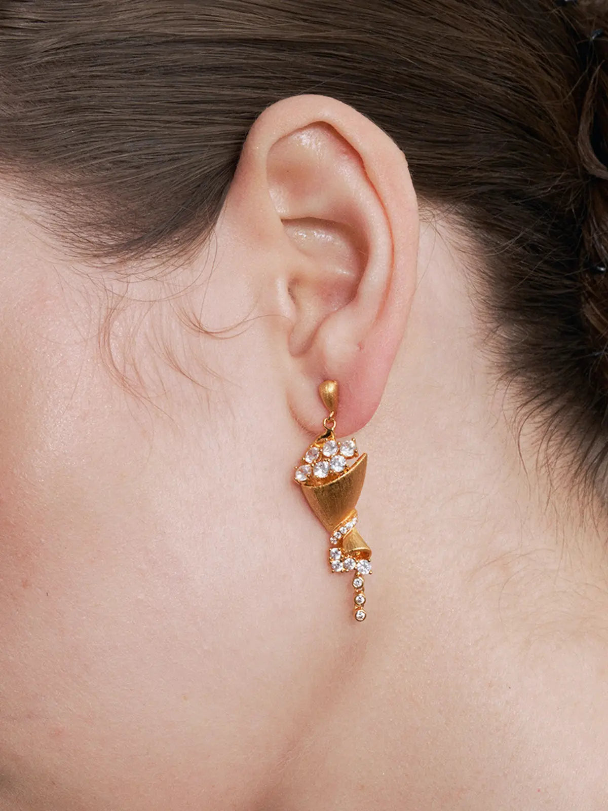 Extreme close-up profile of a model's ear wearing the Tactilery Bouquet Stud Earrings, the sculptural matte brushed gold twisted cone bouquet drop with large round zircon bloom cluster at top, pavé zircon spiral ribbon mid-body, and bezel-set zircon chain tail filling the frame in sharp detail