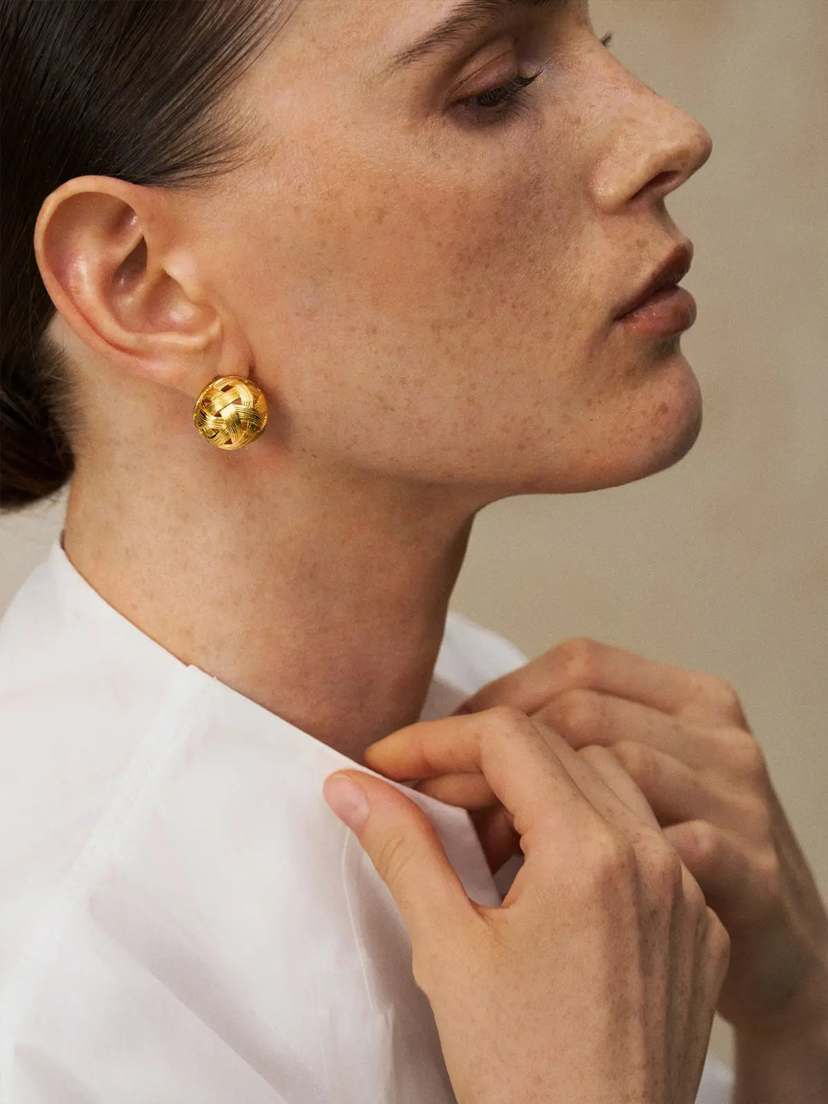 Side profile portrait of a model wearing the Tactilery Brushed Woven Gold Ball Stud Earrings with gaze forward and hands clasped at chest