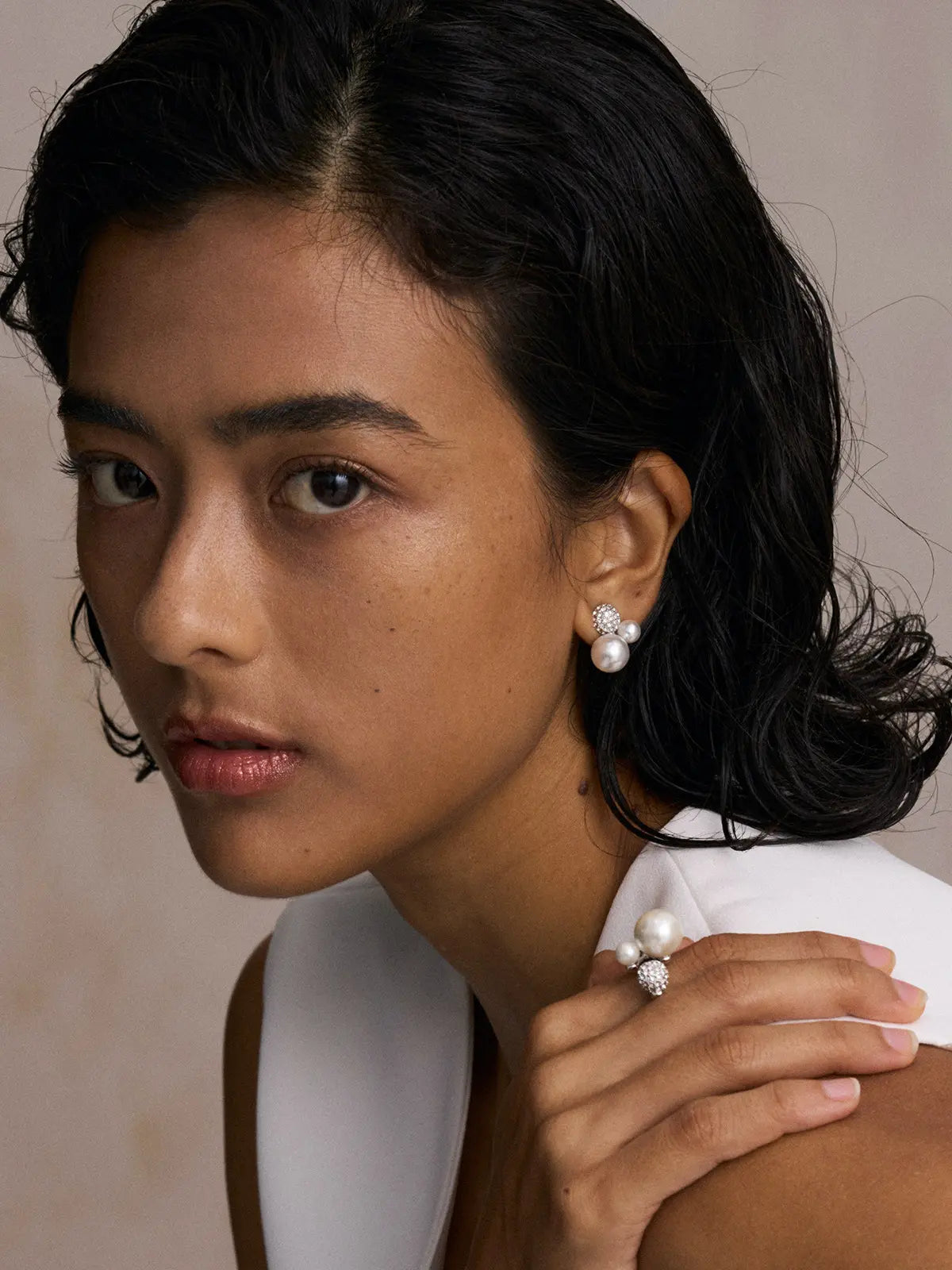 Front-facing editorial portrait of a model wearing the Tactilery Bubble Stud Earrings on both ears and the matching Bubble Ring on the index finger resting on shoulder, the pavé zircon dome sphere with white pearl cluster visible at both ear and finger, against a warm beige background wearing a white sleeveless top