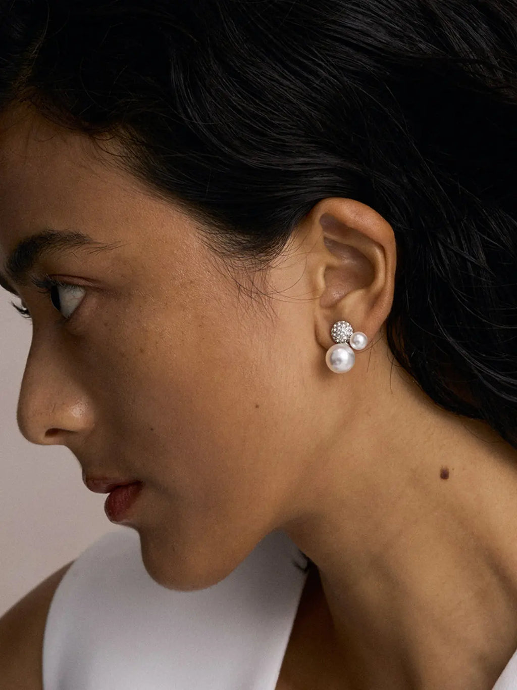 Close-up side profile of a model's ear wearing the Tactilery Bubble Stud Earrings, the pavé zircon dome sphere at top with small and large white pearl spheres clustered below sitting at the lobe, against a warm beige background wearing a white sleeveless top with short dark hair