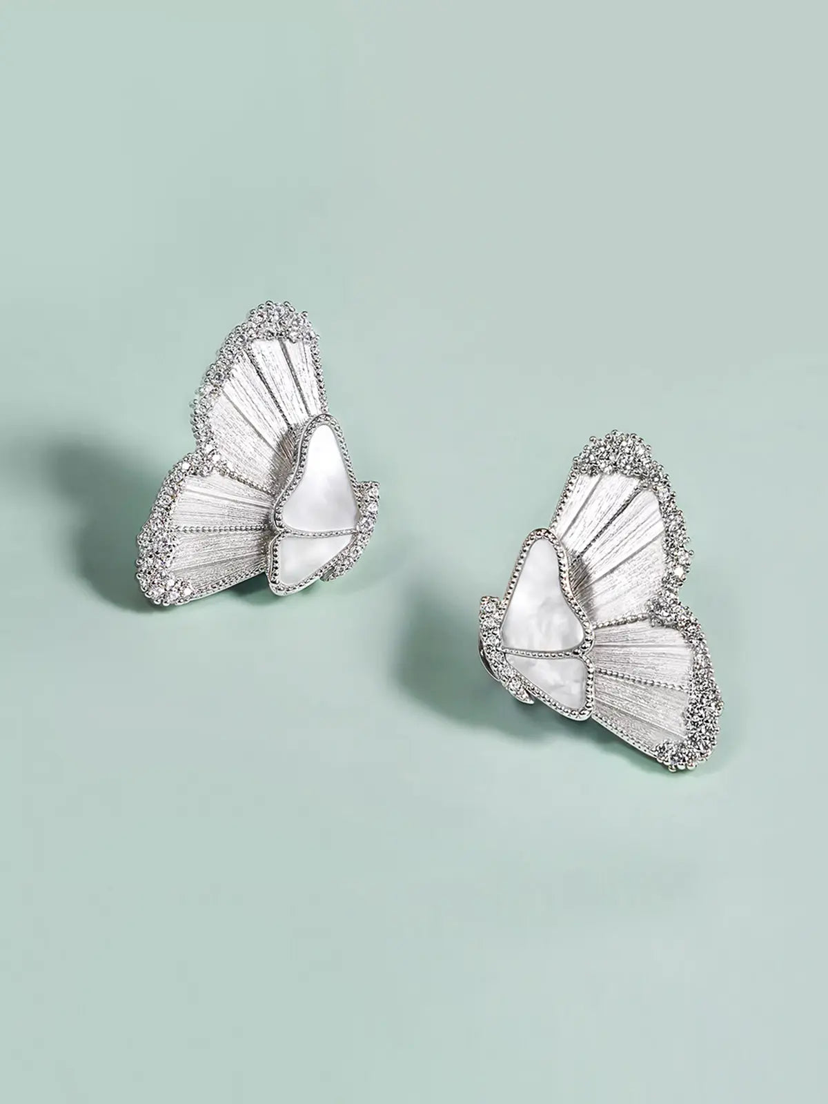 Pair of Tactilery Butterfly Wing Earrings in silver displayed at an angle on a sage green surface, both showing the three-dimensional depth of the sculptural half-butterfly wing form with translucent white mother-of-pearl shell wing panels divided by fine silver wire veins and fully pavé zircon border edging, with silver post pins visible at the body center