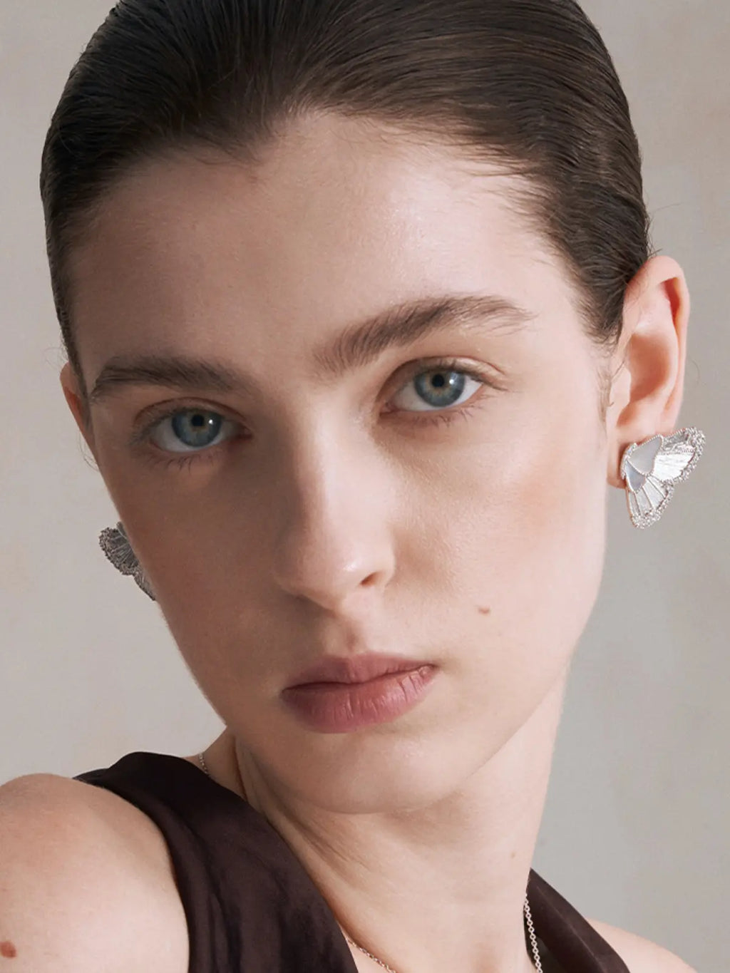 Front-facing editorial portrait of a model wearing the Tactilery Butterfly Wing Earrings on both ears with direct gaze, the large sculptural half-butterfly wing studs with translucent white mother-of-pearl shell wing panels and fully pavé zircon border visible at both lobes, paired with a delicate silver chain necklace, against a warm grey background wearing a dark brown sleeveless top with dark hair slicked back