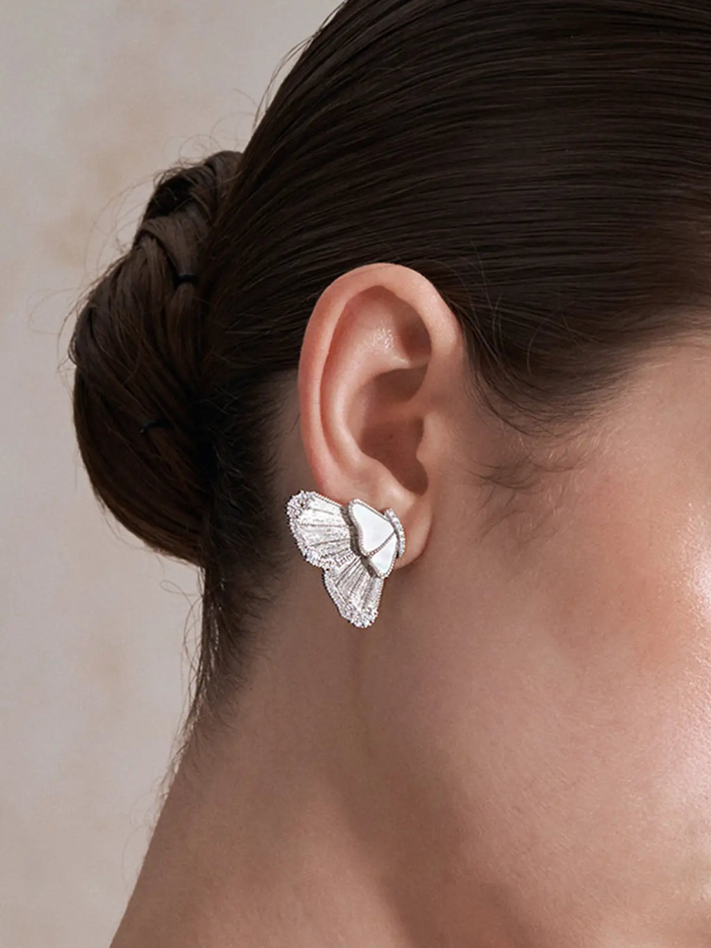 Close-up profile of a model's ear wearing the Tactilery Butterfly Wing Earrings, the large sculptural half-butterfly wing stud with translucent white mother-of-pearl shell wing panels, fine silver wire veins, and fully pavé zircon border sitting at the lobe, against a warm beige background with dark hair in a braided bun