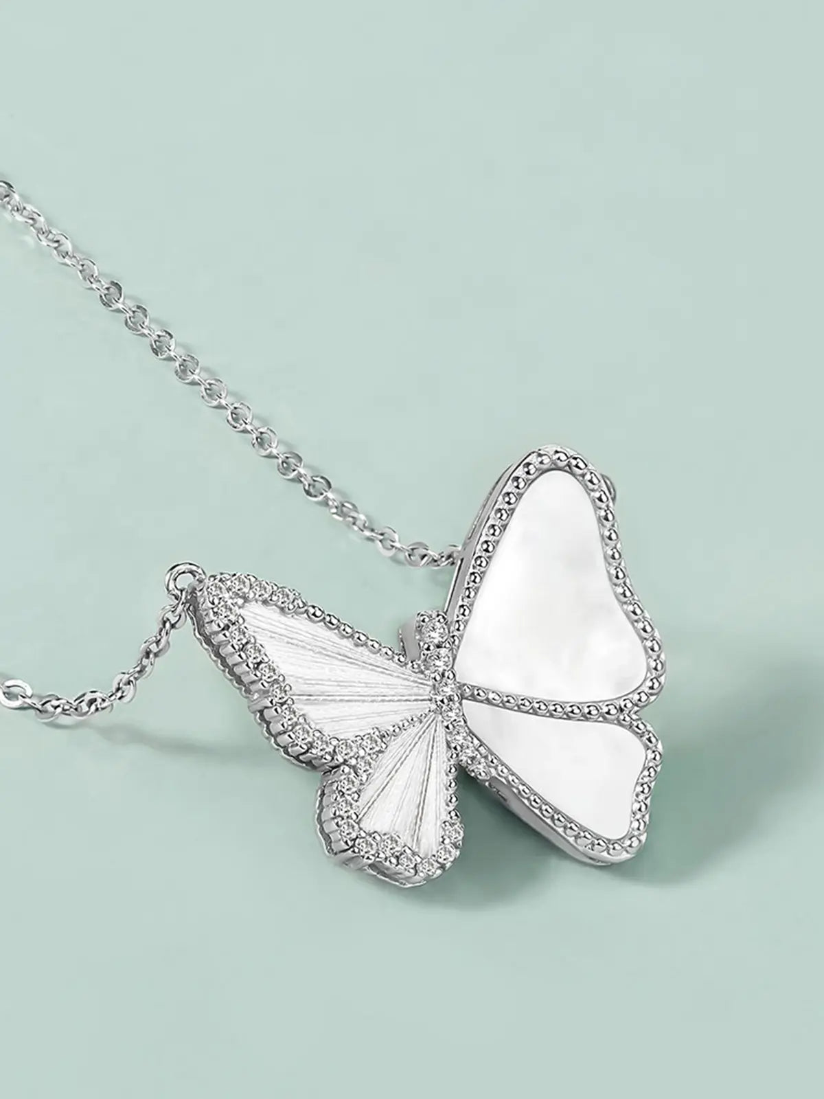Extreme close-up of the Tactilery Butterfly Wing Necklace pendant on a light teal background, the large three-dimensional butterfly shown face-on — the smooth white mother-of-pearl upper wing panels within polished silver pavé zircon-edged frames, the lower wings with engraved silver vein line dividing the mother-of-pearl panel, and the central pavé zircon body column in sharp detail — with the fine polished silver cable chain bail at top