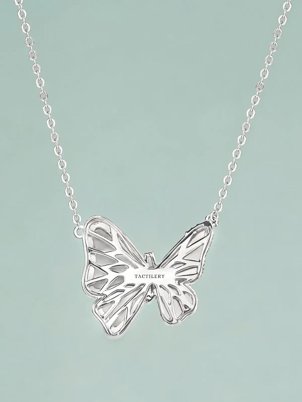 Close-up of the Tactilery Butterfly Wing Necklace pendant on a light teal background at a three-quarter angle, the large three-dimensional butterfly with white mother-of-pearl upper and lower wing panels, continuous pavé zircon wing borders, and central pavé zircon body column shown in detail, with the fine polished silver cable chain and bail visible above