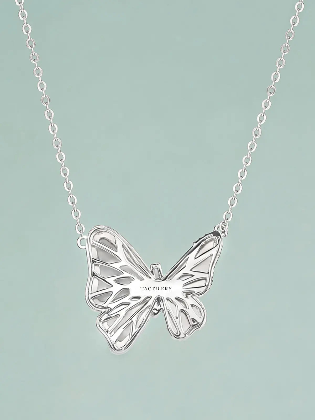 Close-up of the Tactilery Butterfly Wing Necklace pendant on a light teal background at a three-quarter angle, the large three-dimensional butterfly with white mother-of-pearl upper and lower wing panels, continuous pavé zircon wing borders, and central pavé zircon body column shown in detail, with the fine polished silver cable chain and bail visible above