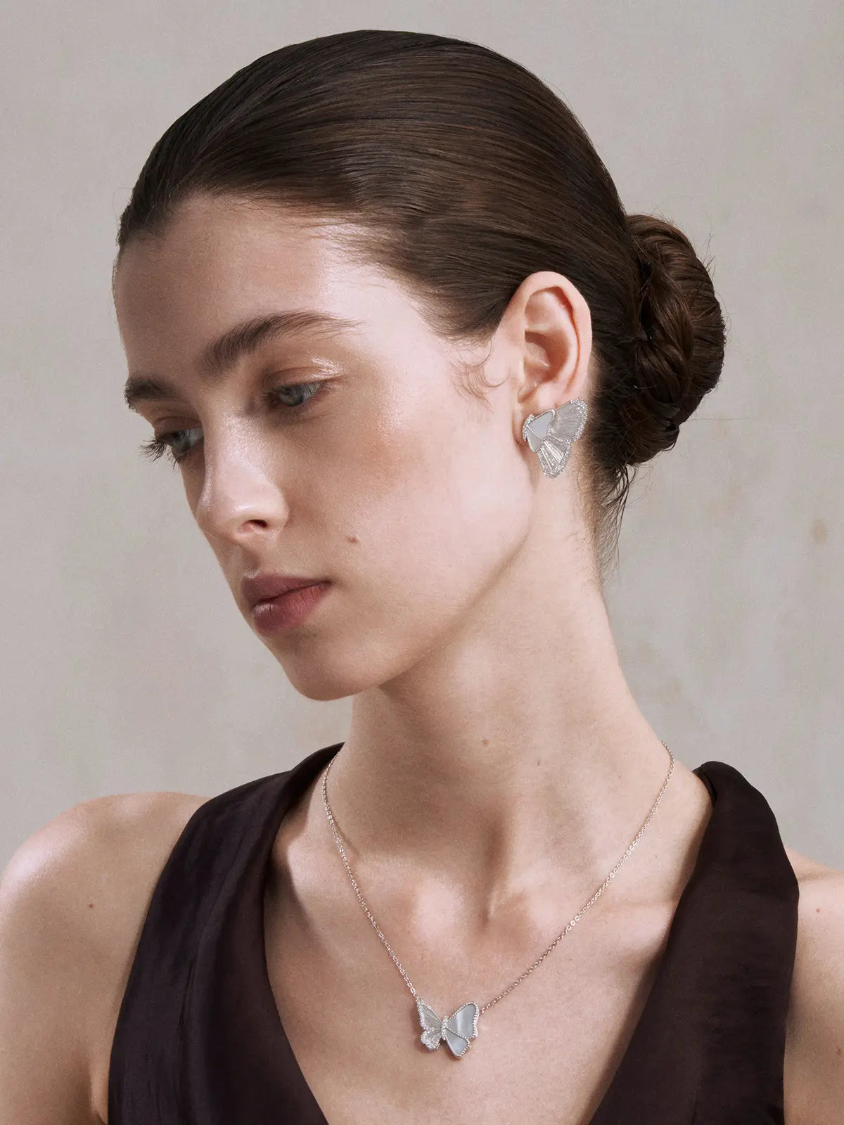Three-quarter portrait of a model wearing the Tactilery Butterfly Wing Necklace with gaze downward, the large three-dimensional butterfly pendant with white mother-of-pearl panel wings edged by pavé zircons and central pavé zircon body visible at the décolletage on a fine polished silver cable chain, against a warm taupe background wearing a white top with dark hair pulled back