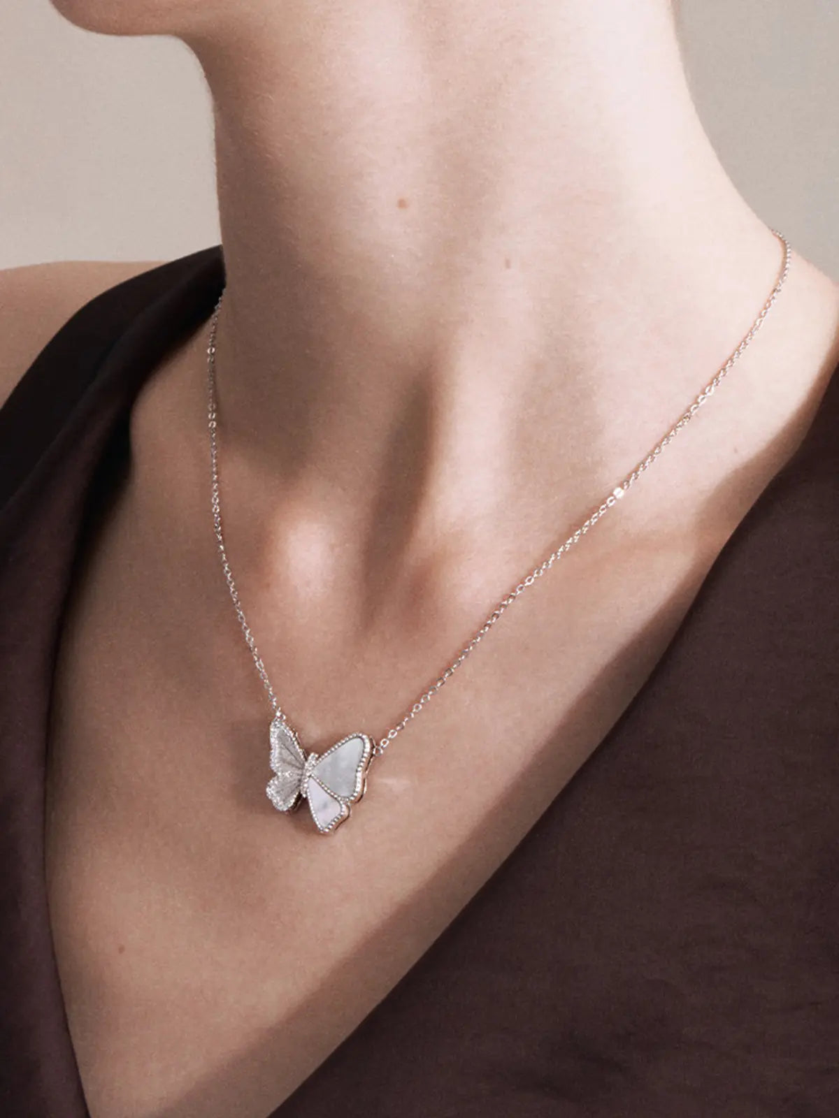 Close-up of a model's décolletage wearing the Tactilery Butterfly Wing Necklace, the large three-dimensional butterfly pendant with white mother-of-pearl panel wings edged by pavé zircons and central pavé zircon body hanging at the neckline on a fine polished silver cable chain, against a warm taupe background wearing a white top
