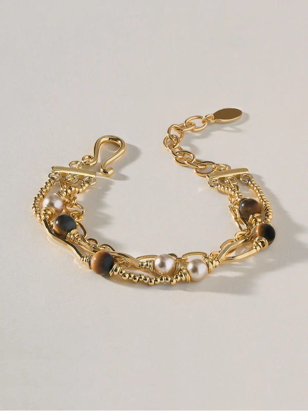 Tactilery Caramel Tiger Eye Multi-Layer Bracelet in polished gold on a warm cream background, the three-strand layered bracelet shown worn and spread open, displaying the fine polished gold beaded ball chain with wire-wrapped caramel brown tiger eye sphere beads, the polished gold oval paperclip link chain with wire-wrapped white freshwater pearl beads, and the polished gold twisted rope chain, with the polished gold S-hook clasp and oval tag charm extension chain at left and the toggle bar clasp at right