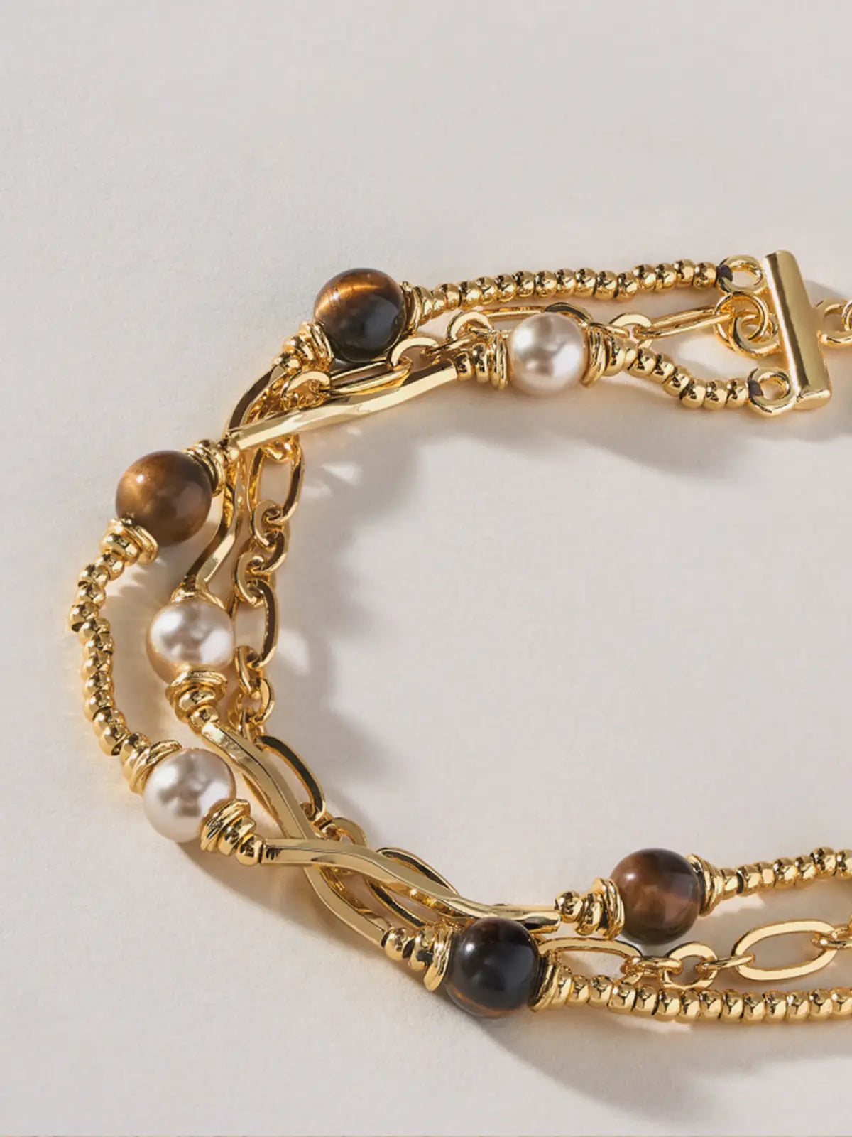 Extreme close-up of the Tactilery Caramel Tiger Eye Multi-Layer Bracelet on a warm cream background, showing the three layered strands in detail — the fine polished gold beaded ball chain with wire-wrapped caramel brown tiger eye sphere beads, the polished gold oval paperclip link chain with wire-wrapped white freshwater pearl beads, and the polished gold twisted rope chain — with the toggle bar clasp visible at top right