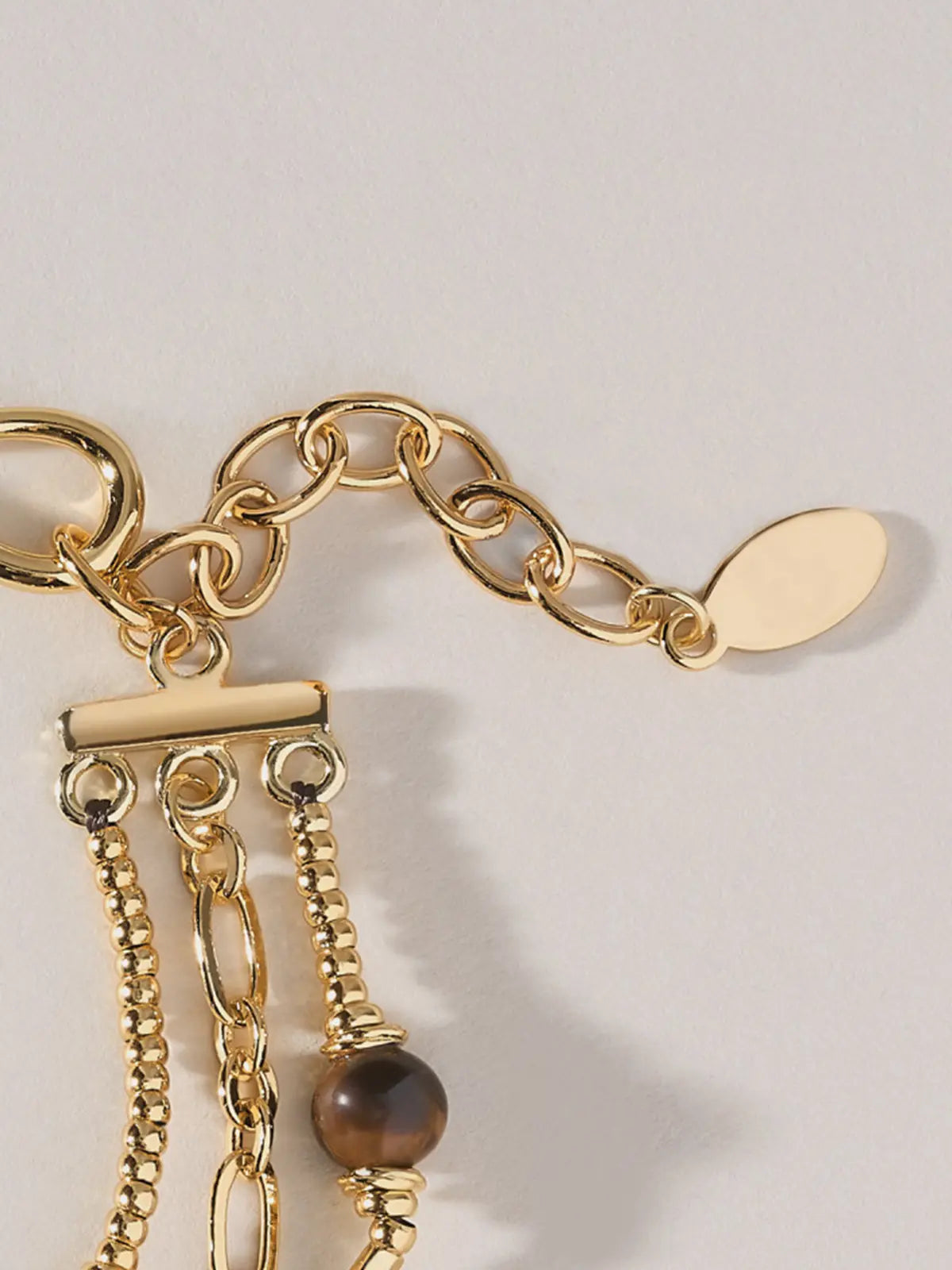Extreme close-up of the Tactilery Caramel Tiger Eye Multi-Layer Bracelet clasp end on a warm cream background, showing the polished gold toggle bar clasp with all three strands — the fine beaded ball chain, the oval paperclip link chain, and the twisted rope chain — fanning out from the bar, and the polished gold S-hook clasp with oval tag charm and multi-link extension chain at top