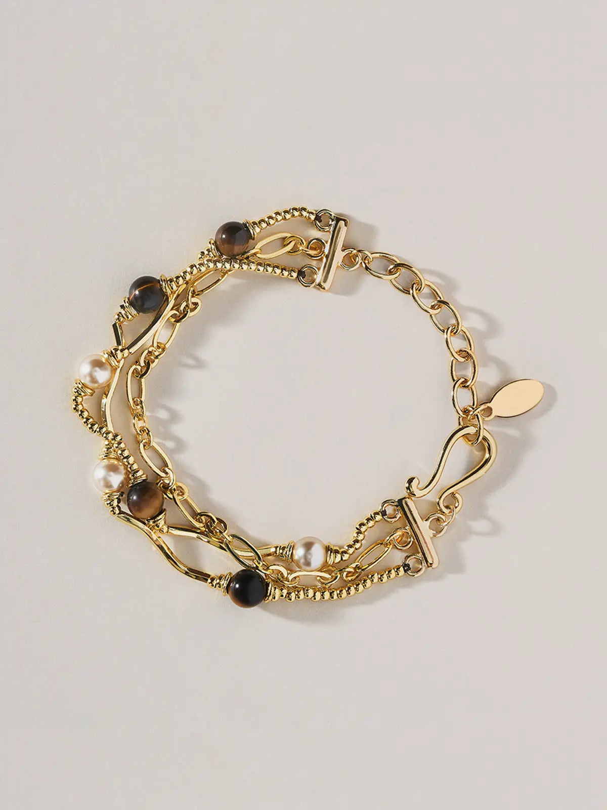 A Tactilery caramel tiger eye multi layer bracelet featuring chain links, rope textures, faux pearls, dark brown beads, and a small oval gold tag, displayed on a light gray background.
