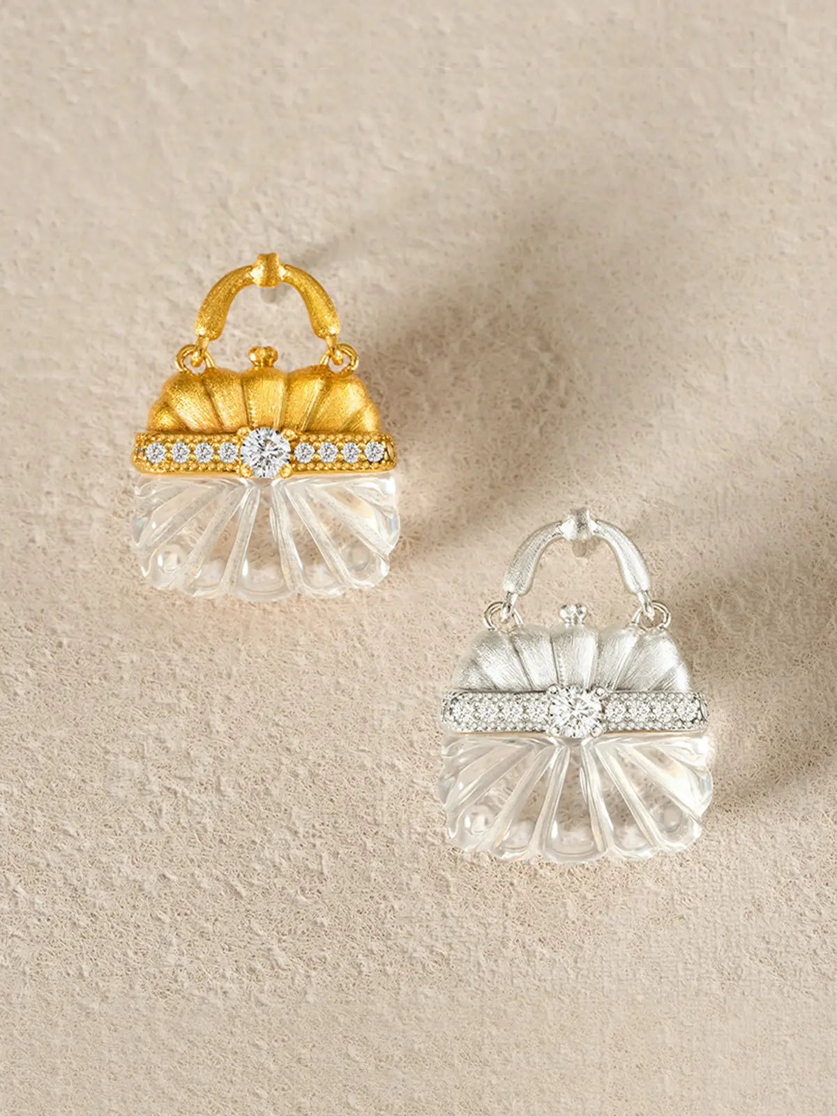Pair of Tactilery Evening Crystal Pouch Earrings displayed face-up on a warm beige linen surface showing both gold and silver colorways side by side — gold colorway with matte brushed gold ribbed dome upper body and pavé zircon clasp band at left, silver colorway with brushed silver ribbed dome upper body and fully pavé zircon clasp band at right — both with carved clear crystal ribbed lower bodies