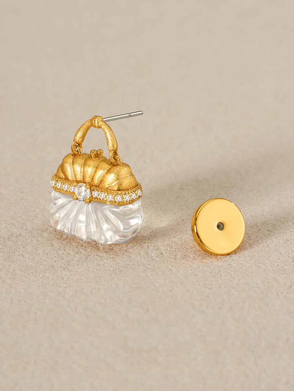 Single Tactilery Evening Crystal Pouch Earring in gold colorway displayed upright on a warm beige linen surface with the gold disc butterfly back removed and placed beside it, showing the silver post pin on the gold loop handle top and the full profile of the miniature handbag form with matte brushed gold ribbed dome upper body, pavé zircon clasp band, and carved clear crystal ribbed lower body