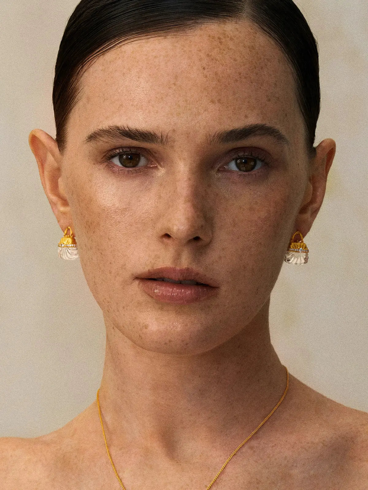 Front-facing editorial portrait of a model wearing the Tactilery Evening Crystal Pouch Earrings in gold colorway on both ears, the miniature handbag stud with matte brushed gold ribbed dome upper body, pavé zircon clasp band, and carved clear crystal ribbed lower body sitting at the lobe, paired with a delicate gold cable chain necklace, against a warm beige background with hair slicked back