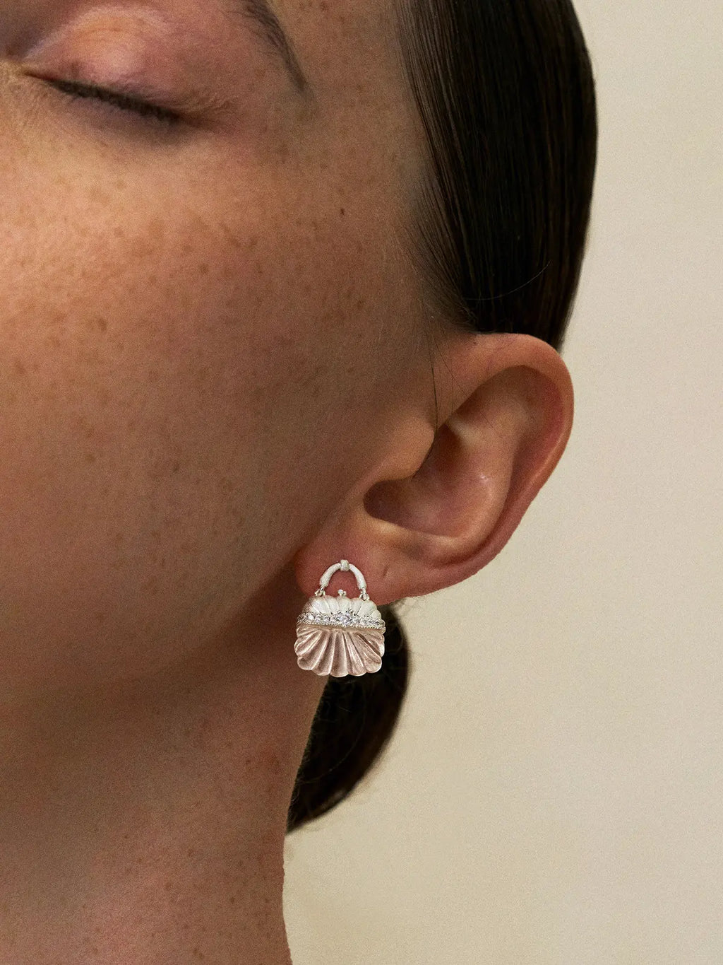 Close-up profile of a model's ear wearing the Tactilery Evening Crystal Pouch Earrings in silver colorway, the miniature handbag stud with brushed silver ribbed dome upper body, fully pavé zircon clasp band with central prong-set round zircon, and carved clear crystal ribbed lower body sitting at the lobe, against a warm cream background