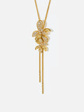Tactilery Flower Narrative Long Necklace in matte brushed gold on a white background, the long Y-drop necklace with a fine polished gold satellite chain suspending a cluster of five sculptural matte brushed gold elements — three dimensional five-petal flower blossoms, small leaf sprigs, and scattered prong-set round zircon accents — gathered at the drop point, with a fine polished gold cable chain extension and lobster clasp