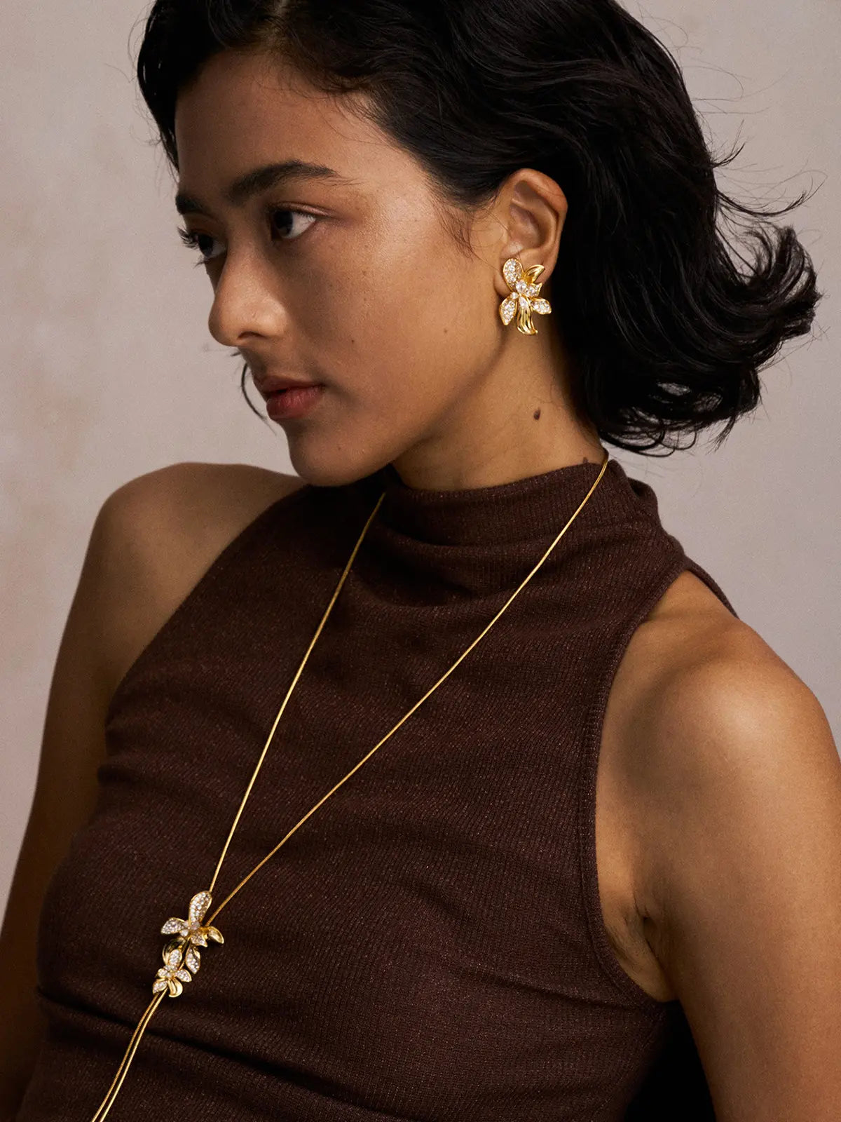Three-quarter portrait of a model wearing the Tactilery Flower Narrative Long Necklace with gaze downward, the long Y-drop with sculptural matte brushed gold flower blossom and leaf cluster with prong-set round zircon accents visible at the sternum on a fine polished gold satellite chain, against a warm taupe background wearing a white sleeveless top with dark hair in a low bun