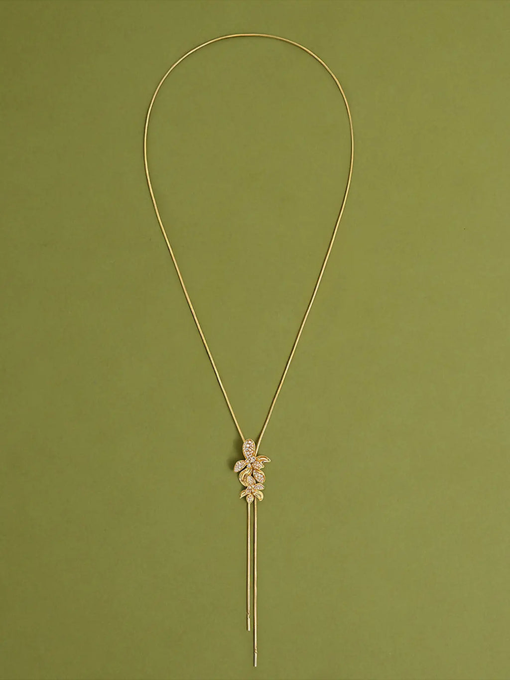 Close-up of a model's torso wearing the Tactilery Flower Narrative Long Necklace, the sculptural matte brushed gold flower blossom and leaf cluster with prong-set round zircon accents shown in detail at the sternum, against a warm taupe background wearing a white sleeveless top