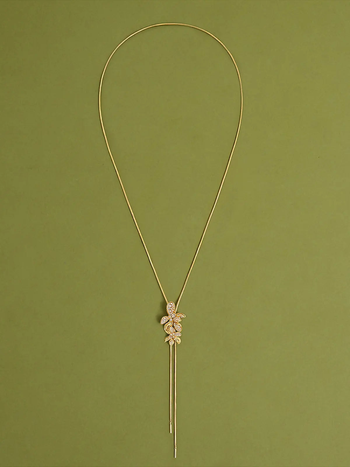 Close-up of a model's torso wearing the Tactilery Flower Narrative Long Necklace, the sculptural matte brushed gold flower blossom and leaf cluster with prong-set round zircon accents shown in detail at the sternum, against a warm taupe background wearing a white sleeveless top