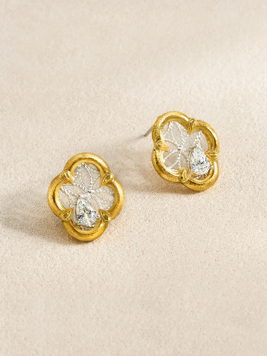 Pair of Tactilery Dream Crabapple Window Earrings displayed at an angle on a warm cream textured surface, one showing the full front face of the quatrefoil stud with matte brushed gold scalloped border, silver twisted wire star lattice, and central pear-cut zircon, one showing the three-dimensional depth with silver post pin visible