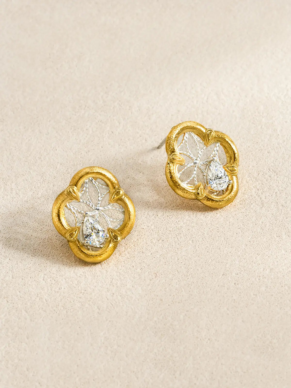 Pair of Tactilery Dream Crabapple Window Earrings displayed at an angle on a warm cream textured surface, one showing the full front face of the quatrefoil stud with matte brushed gold scalloped border, silver twisted wire star lattice, and central pear-cut zircon, one showing the three-dimensional depth with silver post pin visible