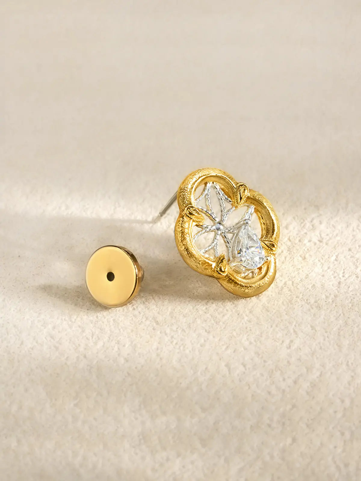 Single Tactilery Dream Crabapple Window Earring displayed upright on a warm cream textured surface with the gold disc butterfly back removed and placed beside it, showing the silver post pin and the full front face of the quatrefoil stud with matte brushed gold scalloped border, silver twisted wire star lattice, and central pear-cut zircon