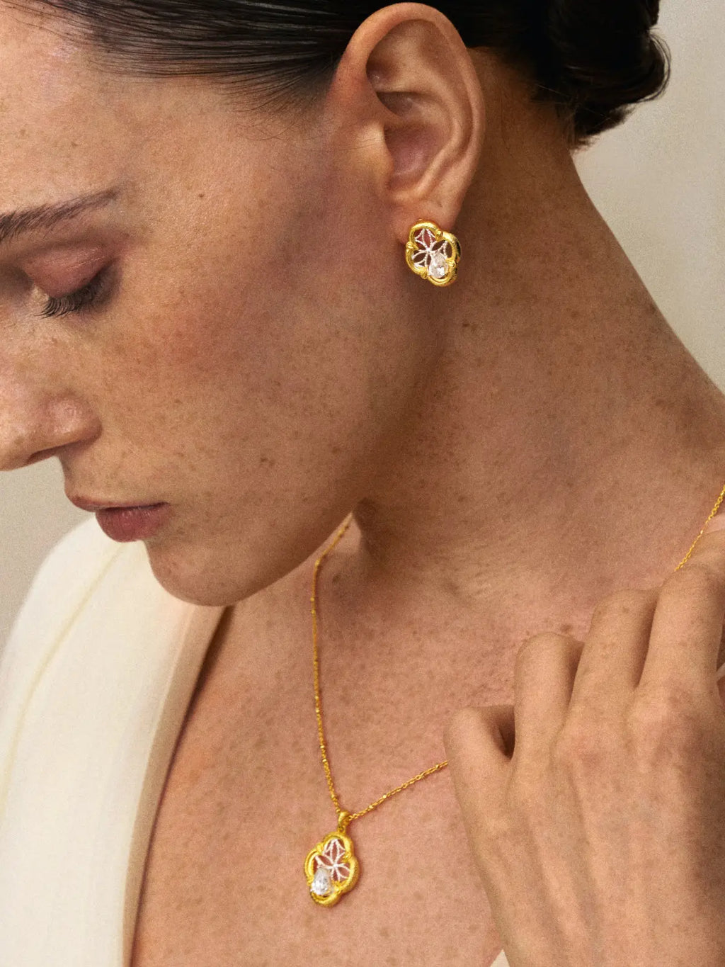 Close-up profile of a model's ear wearing the Tactilery Dream Crabapple Window Earrings with gaze downward, the quatrefoil stud with matte brushed gold scalloped border and silver twisted wire star lattice with central pear-cut zircon at the lobe, paired with a matching Dream Crabapple Window pendant necklace on a gold chain, against a warm beige background wearing a white draped top