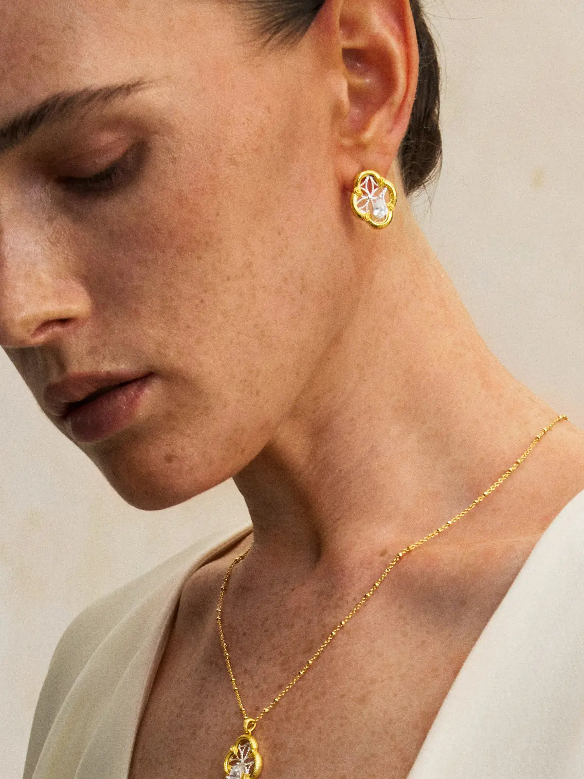 Three-quarter portrait of a model wearing the Tactilery Dream Crabapple Window Earrings with gaze downward, the quatrefoil stud with matte brushed gold scalloped border and silver twisted wire star lattice with central pear-cut zircon visible at the lobe, paired with a matching Dream Crabapple Window pendant necklace on a gold chain, against a warm beige background wearing a white draped top