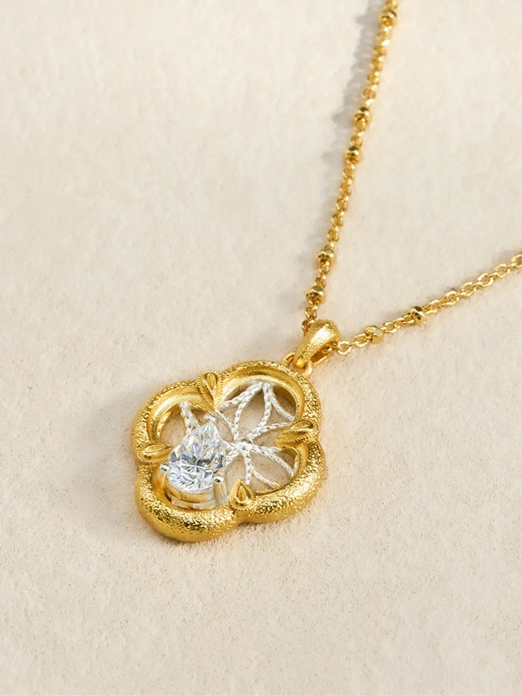Close-up of the Tactilery Dream Crabapple Window Necklace pendant displayed on a warm cream textured surface at an angle, the open-frame four-lobe crabapple flower pendant with wide matte sand-textured gold border ring, small teardrop bud accents at the four cardinal points, fine silver wire star-web of eight radiating twisted threads, and large prong-set pear-cut zircon at lower center, with the polished gold satellite chain and bail visible above