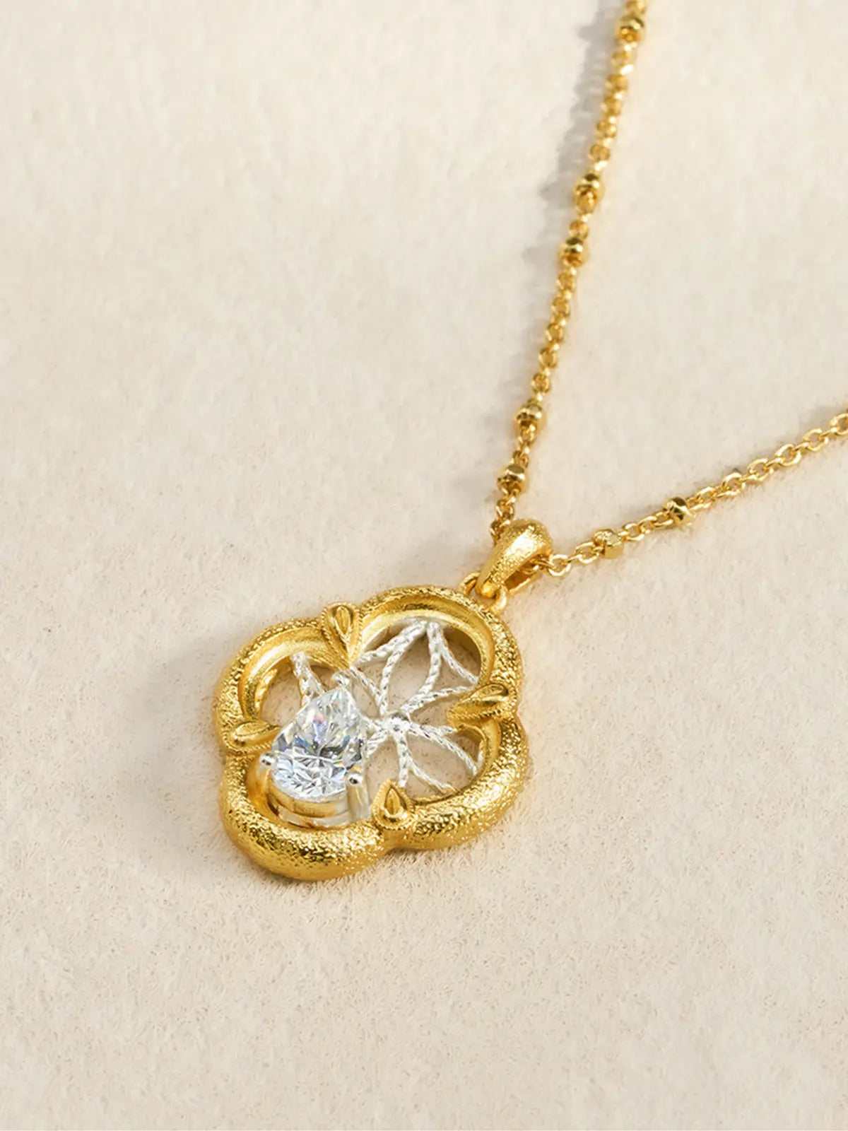 Close-up of the Tactilery Dream Crabapple Window Necklace pendant displayed on a warm cream textured surface at an angle, the open-frame four-lobe crabapple flower pendant with wide matte sand-textured gold border ring, small teardrop bud accents at the four cardinal points, fine silver wire star-web of eight radiating twisted threads, and large prong-set pear-cut zircon at lower center, with the polished gold satellite chain and bail visible above