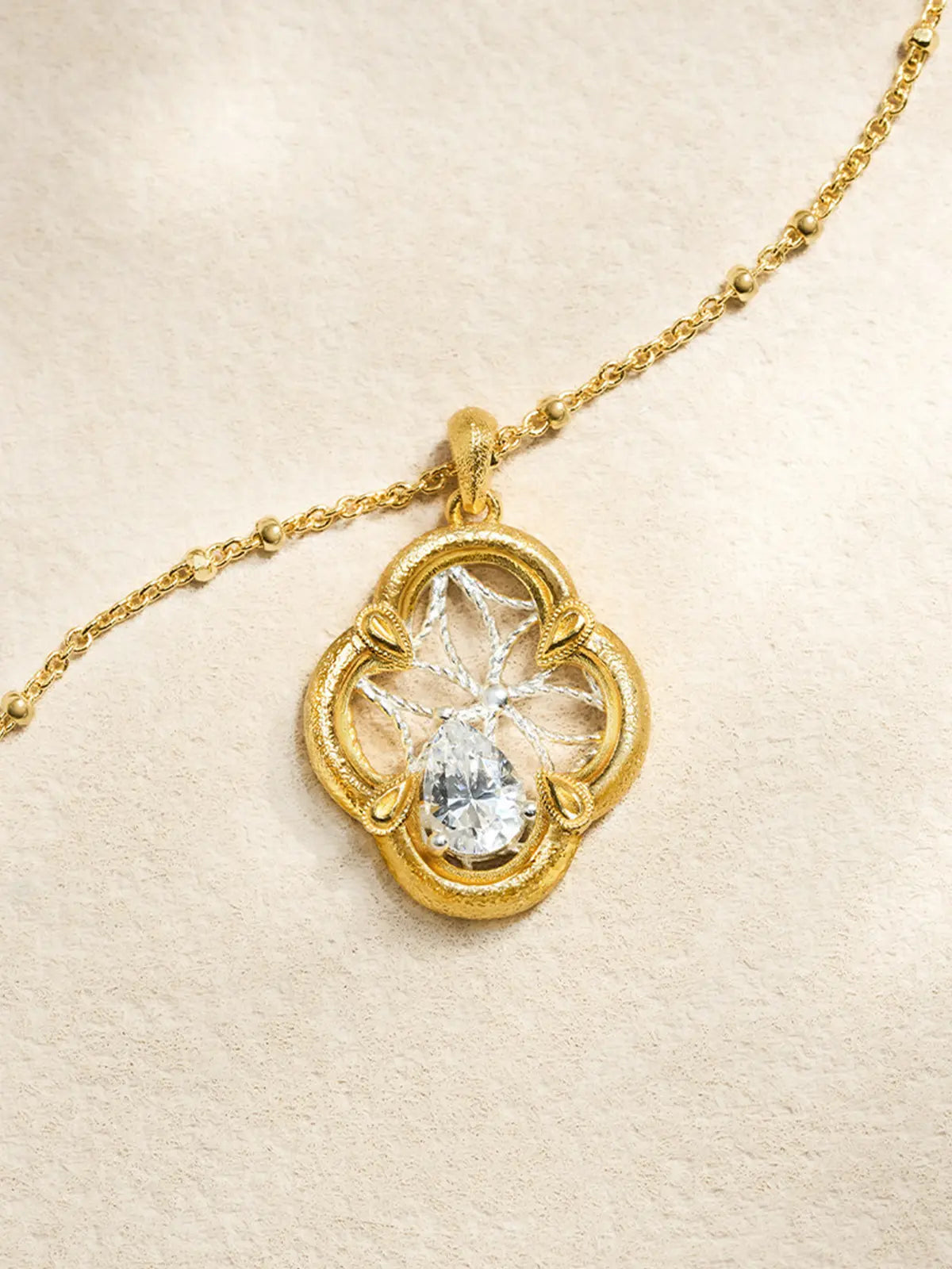 Close-up of the Tactilery Dream Crabapple Window Necklace pendant displayed on a warm cream textured surface showing the back of the pendant, the smooth matte sand-textured gold back face of the four-lobe crabapple flower border ring with the fine silver wire star-web threads visible through the open frame from behind, and the large prong-set pear-cut zircon prong setting visible at lower center, with the polished gold satellite chain bail at top