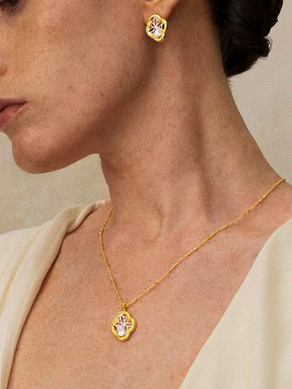Side profile portrait of a model wearing the Tactilery Dream Crabapple Window Necklace with gaze forward, the open-frame four-lobe crabapple flower pendant with matte sand-textured gold border, fine silver wire star-web interior, and large prong-set pear-cut zircon visible at the décolletage on a polished gold satellite chain, paired with matching dream crabapple window stud earrings visible at the ear, against a warm taupe background wearing a white draped top with dark hair in a low bun